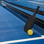 Pickleball paddle resting against net with yellow ball on blue court